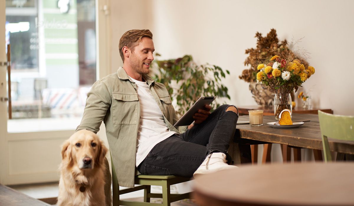 A man sits in a cozy cafe, smiling while using a tablet, with a golden retriever by his side. A table holds coffee, cake, and flowers. Relaxed atmosphere.